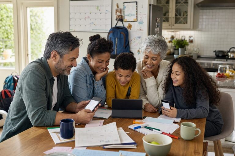 A multigenerational family around a kitchen table coordinating schedules on phones, with a paper calendar and backpacks in view.